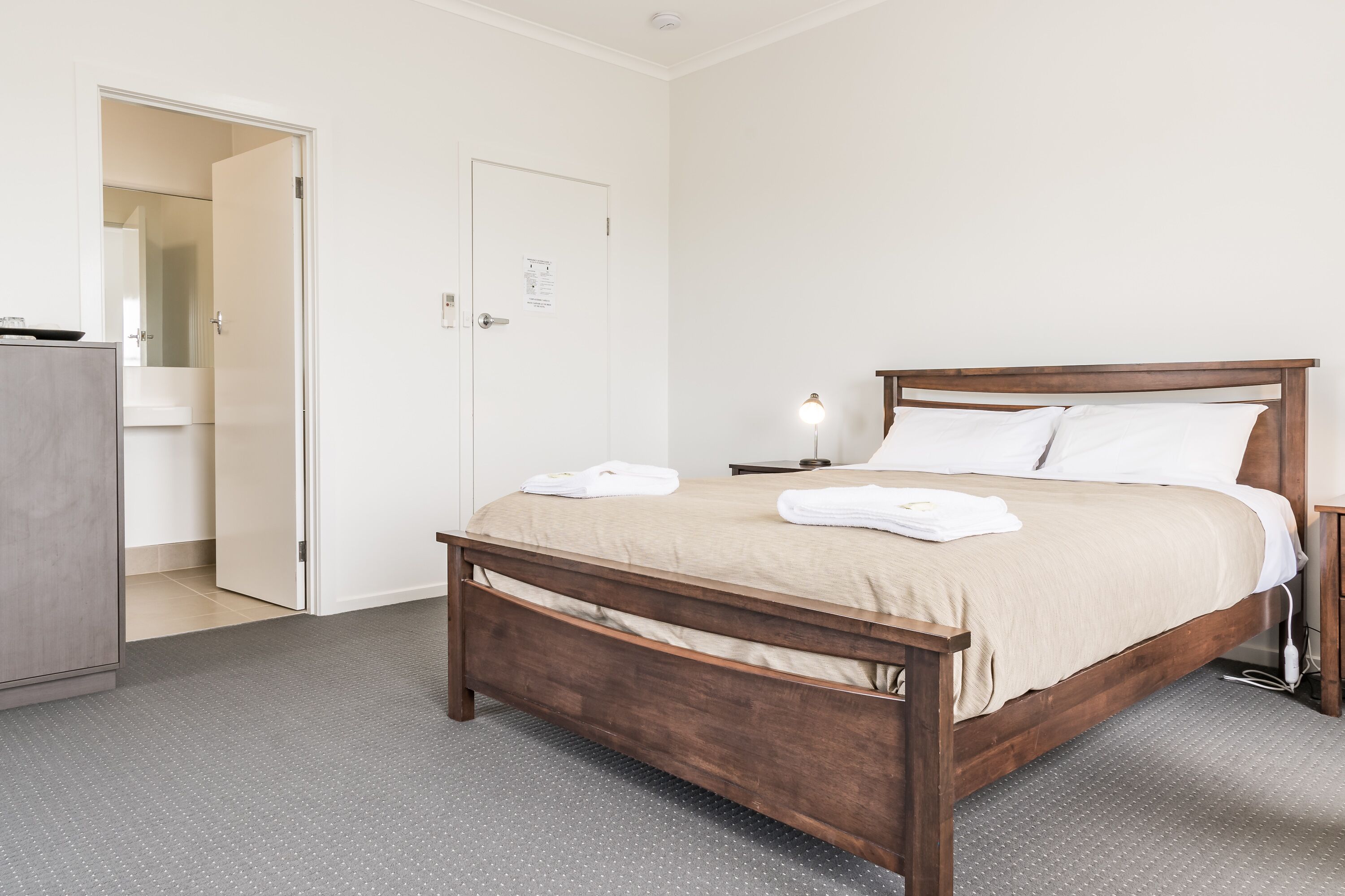 Un lit bien fait, avec un cadre en bois et une couvre-lit beige, une table de chevet avec une lampe, et une porte donnant sur une salle de bain.