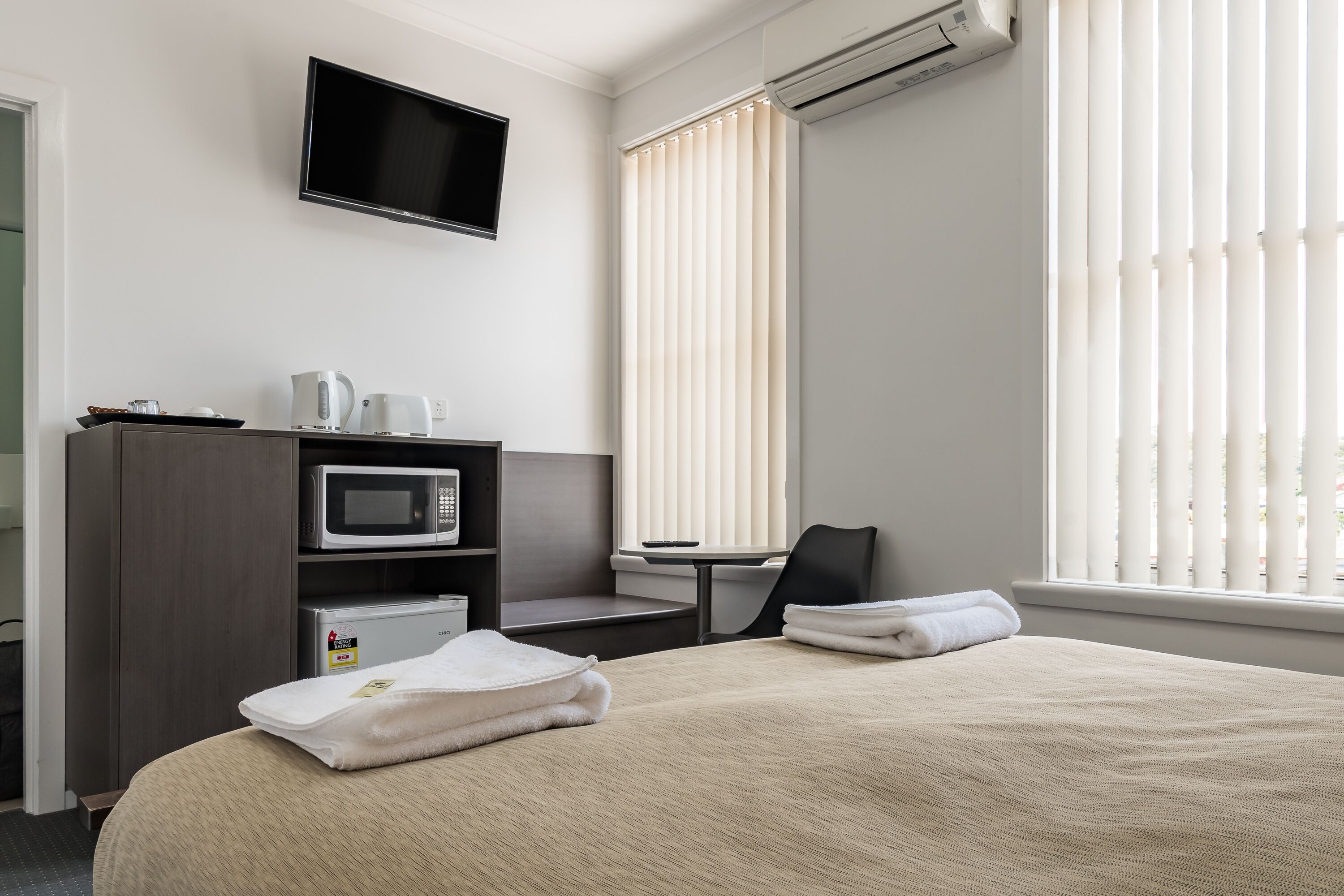 A hotel room with a bed, a desk, a microwave, a TV, and a window with blinds.