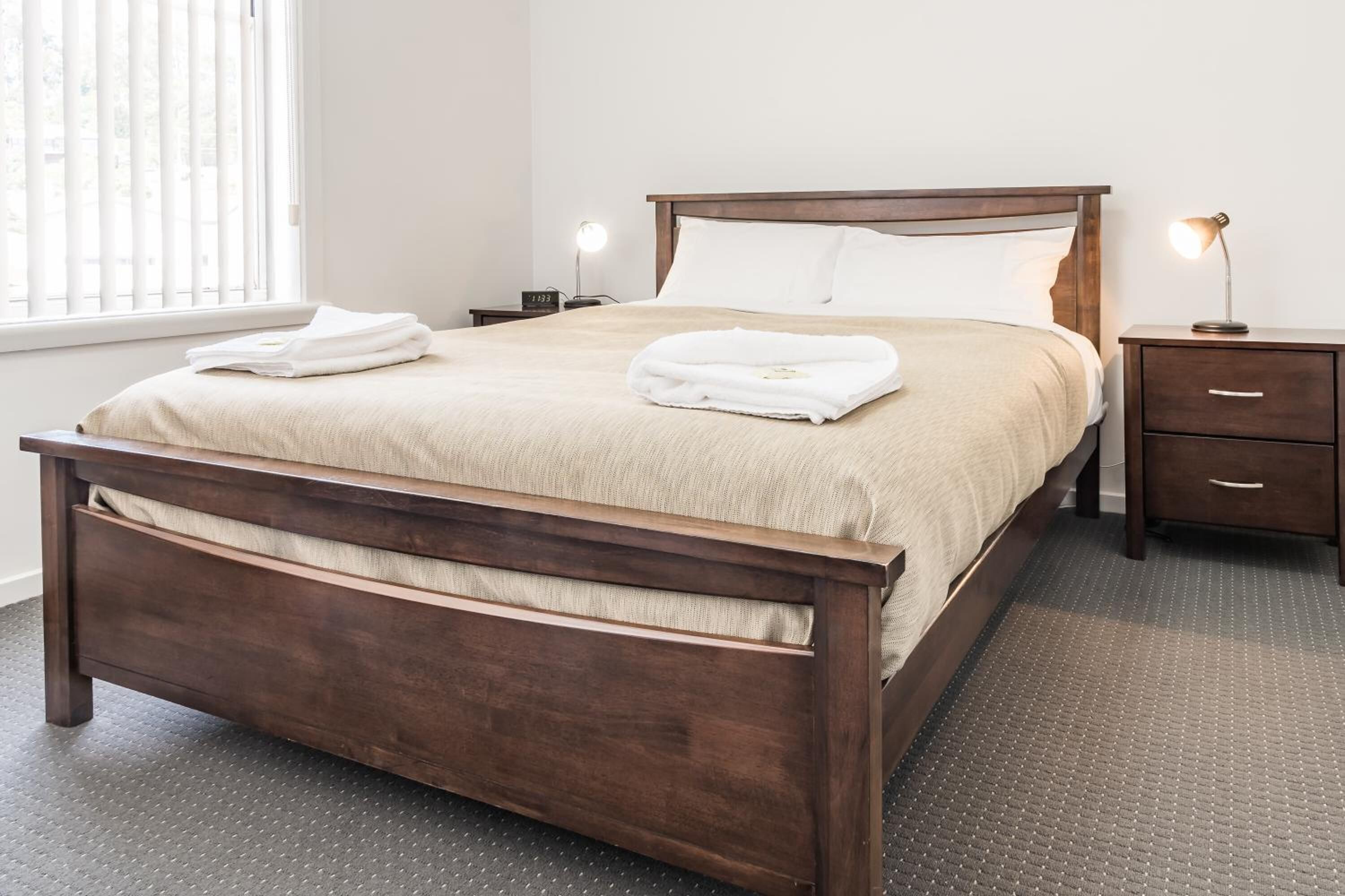 A neatly made bed with a wooden frame and a beige bedspread, accompanied by folded towels. There is a bedside table with a lamp and a small drawer.