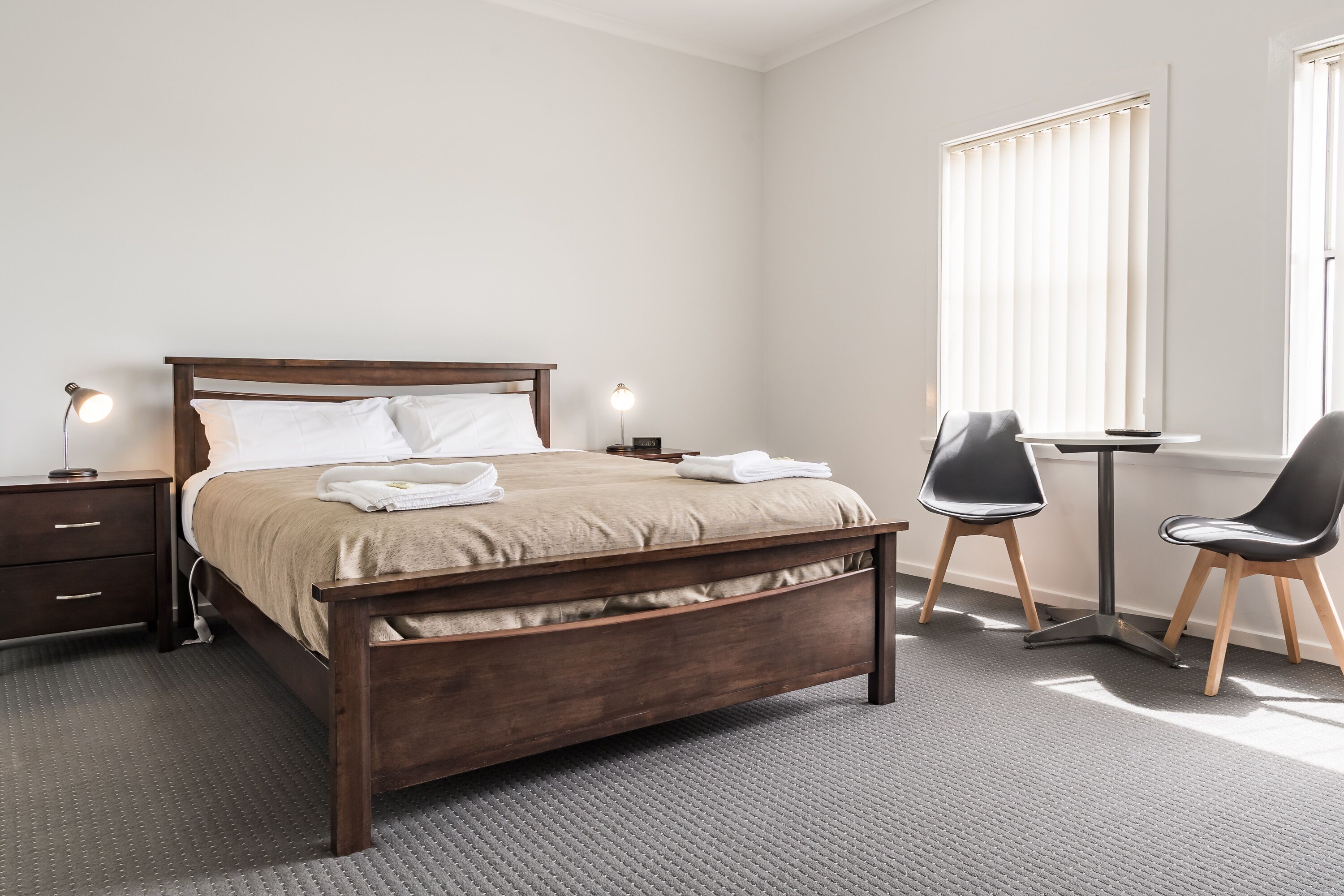 Un dormitorio ordenado con cama de madera, mesitas de noche, un escritorio y sillas.
