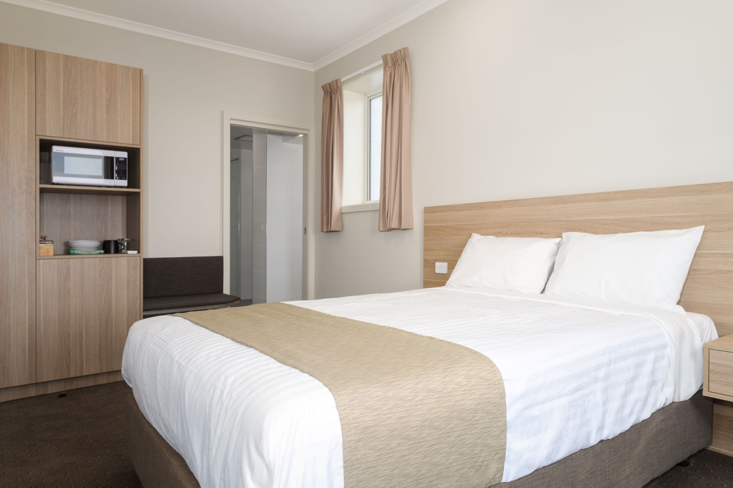 A neatly arranged hotel room with a bed, a microwave on a shelf, a door, and a window with curtains.