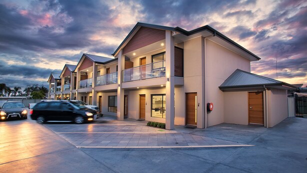 Un edificio residencial moderno de varios pisos con un balcón en el segundo piso, un garaje y un camino de acceso con coches estacionados.