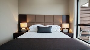 Una cama bien tendida con sábanas blancas y una manta oscura, flanqueada por dos mesitas de noche con lámparas.