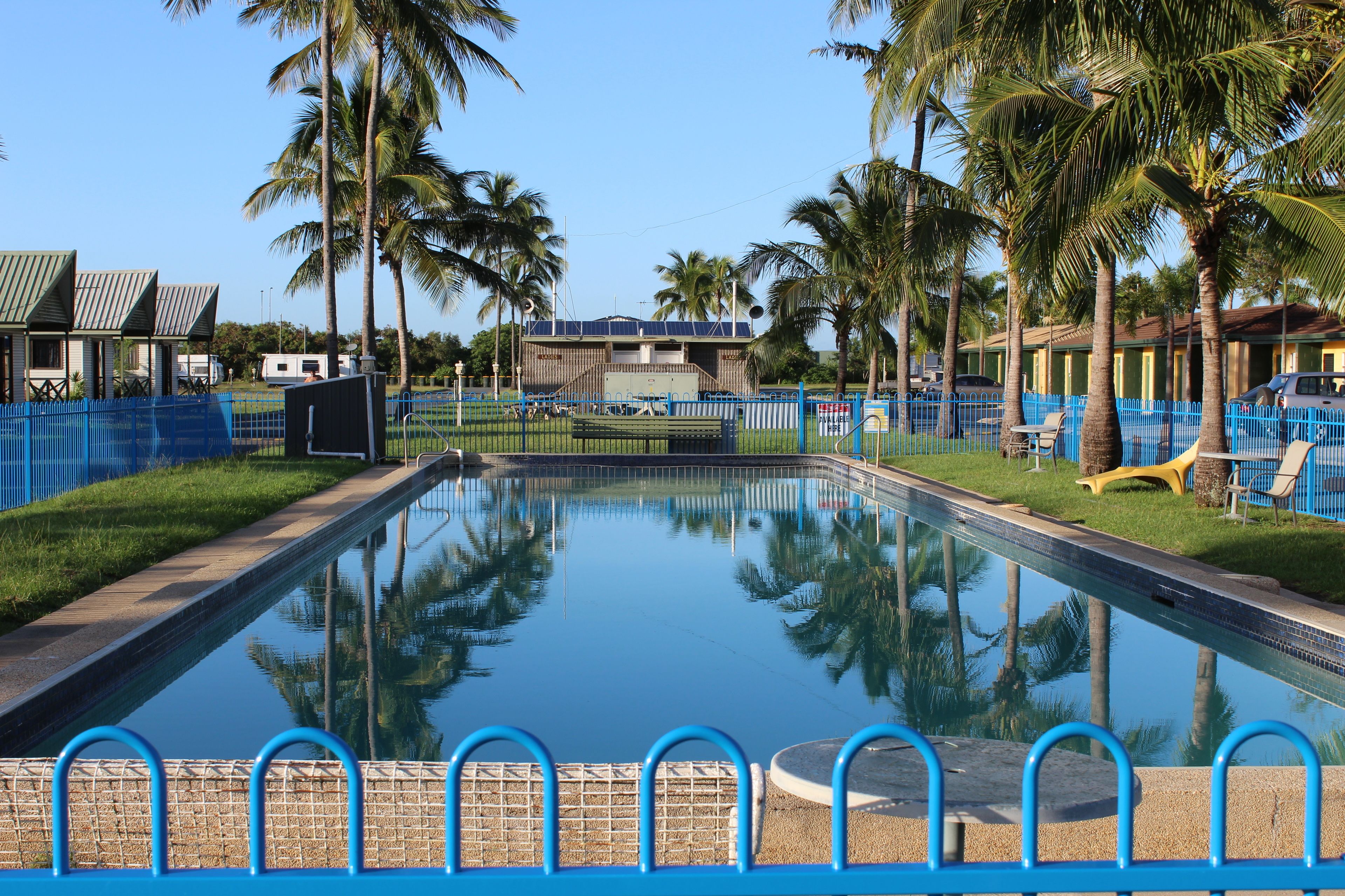 Une piscine rectangulaire entourée d’une clôture bleue, bordée de palmiers et avec un bâtiment au toit bleu en arrière-plan.