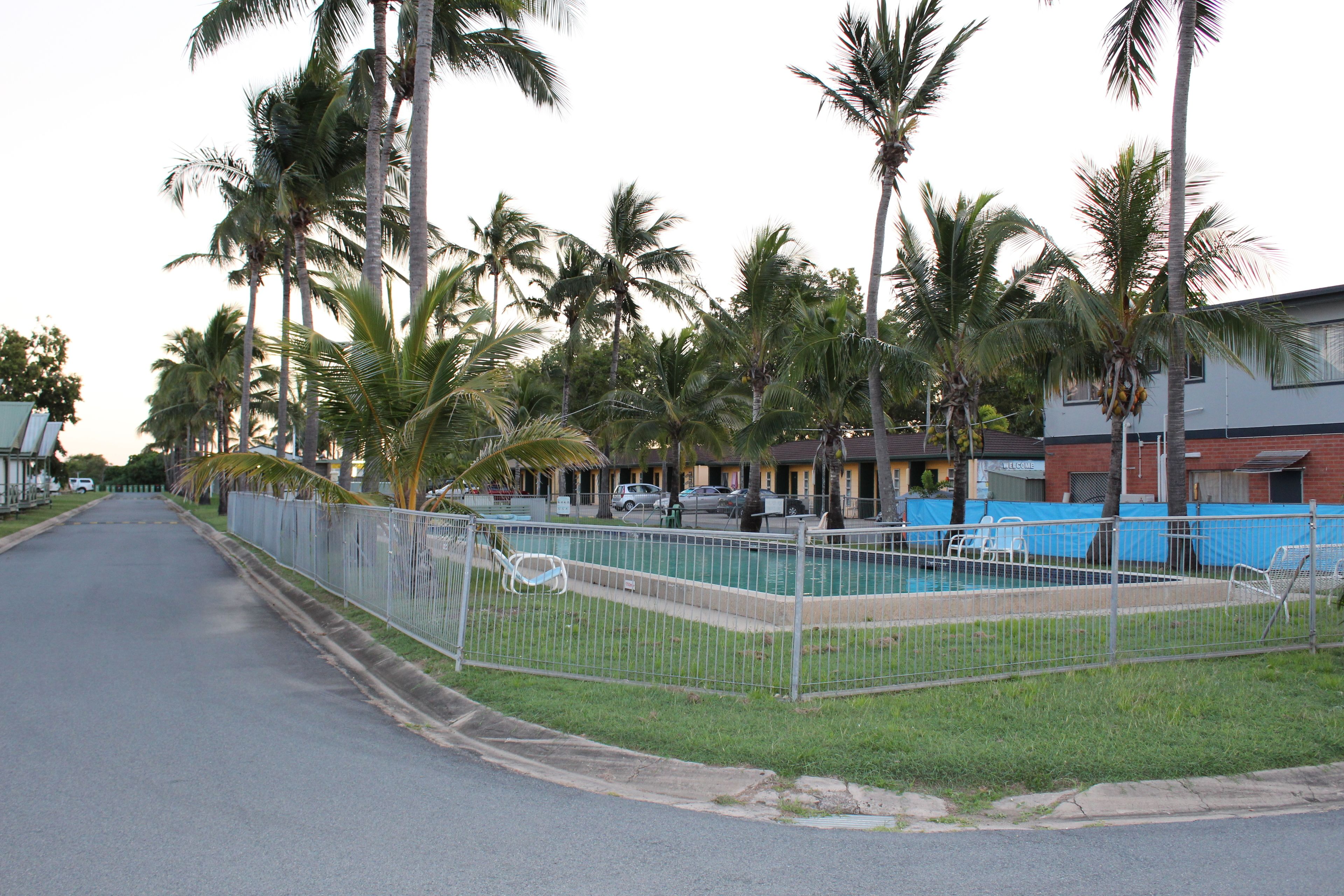 Vista de la calle con un área de piscina cercada, palmeras y un edificio con un letrero de 'Welcome'.