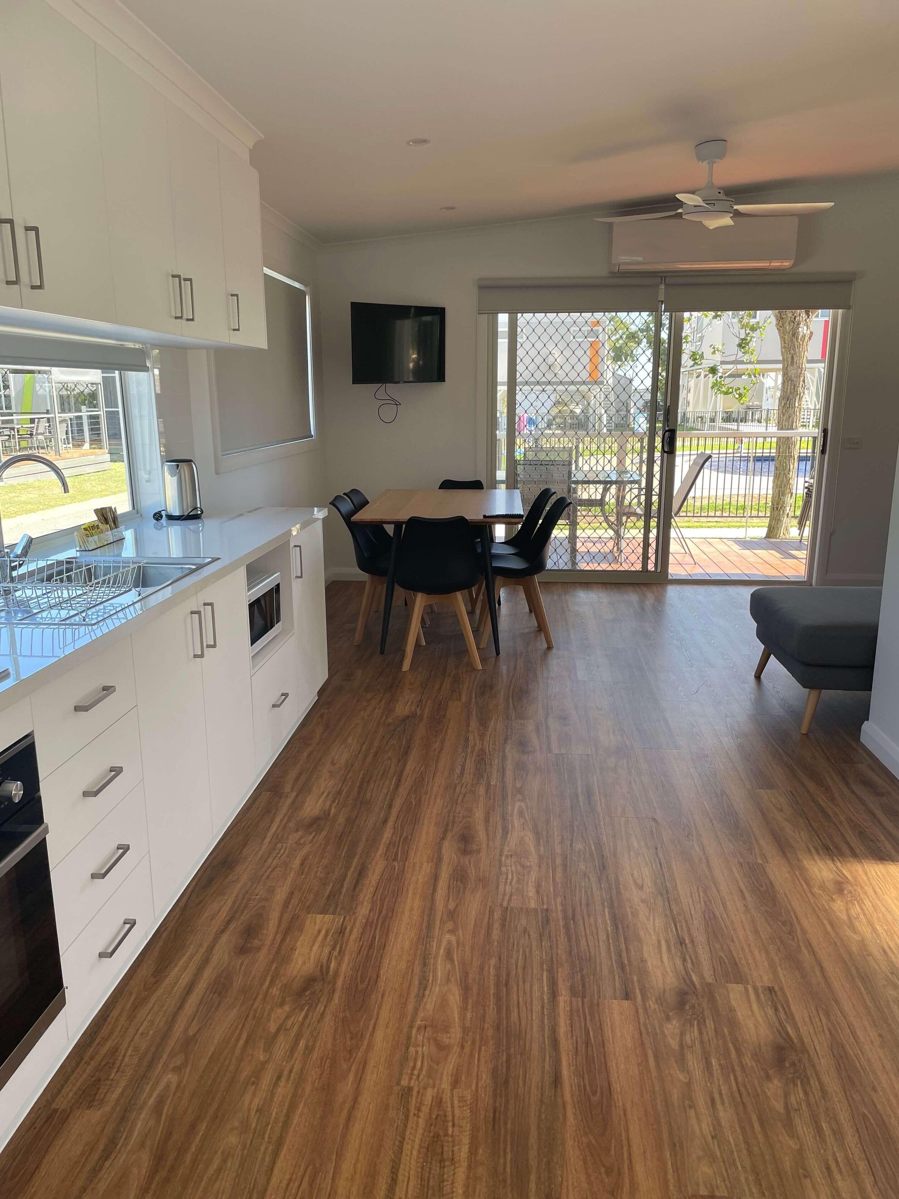 Una cocina moderna con armarios blancos, un comedor con mesa y sillas de madera, y un ventanal corredero que da a un patio exterior.