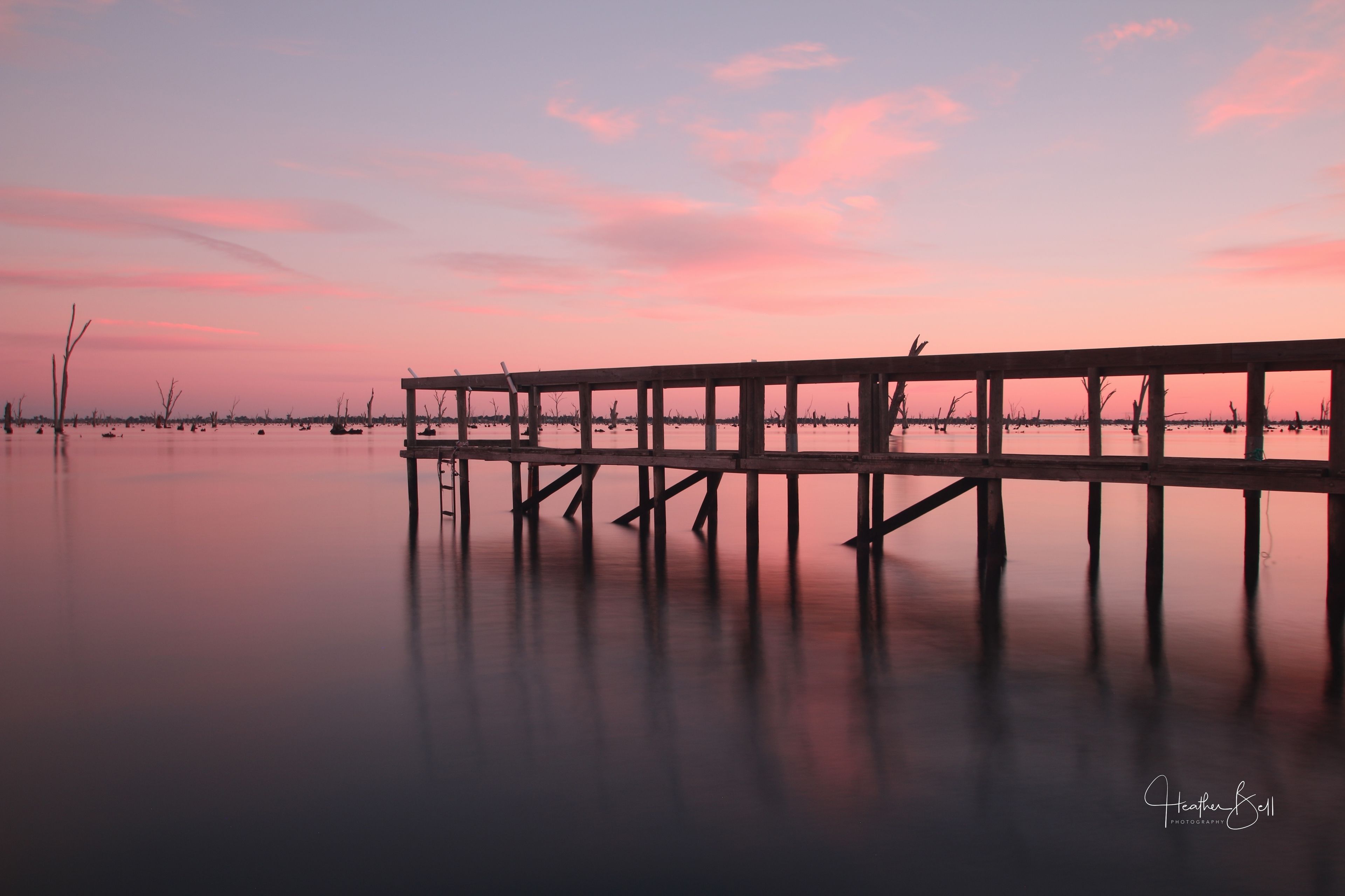 A wooden pier extending into calm water with a pink and purple sky in the background.