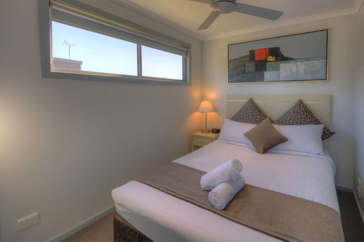 Quarto de hotel com cama, mesa de cabeceira, luminária, ventilador de teto, janela e um quadro na parede.
