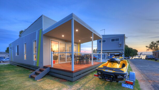 Une maison moderne à deux étages, dotée d’un patio couvert, d’un jet ski jaune et offrant une vue sur l’eau.