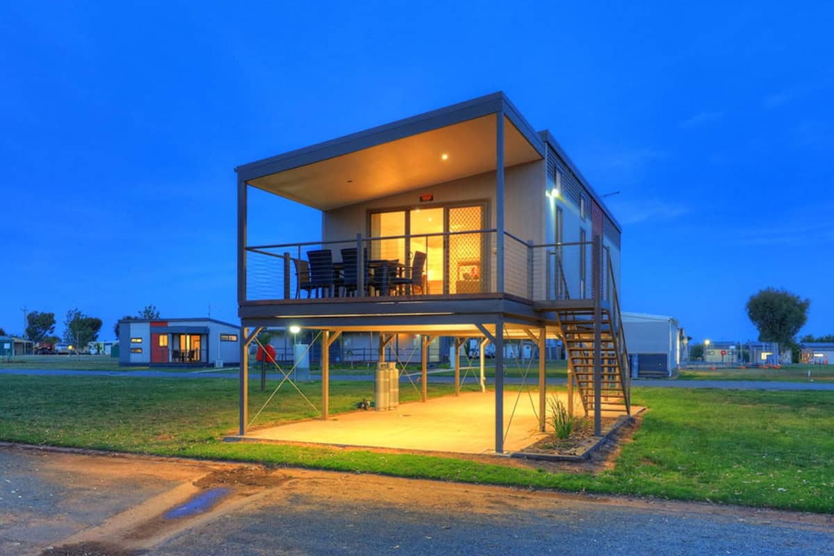 Um edifício moderno de dois andares com varanda e área externa coberta, cercado por outras construções semelhantes.