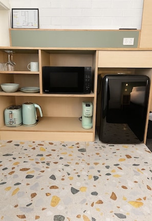 A compact kitchenette with a microwave, coffee maker, and kettle on shelves, a refrigerator, and a terrazzo floor. - Wilsons Promontory Motel (Foster)