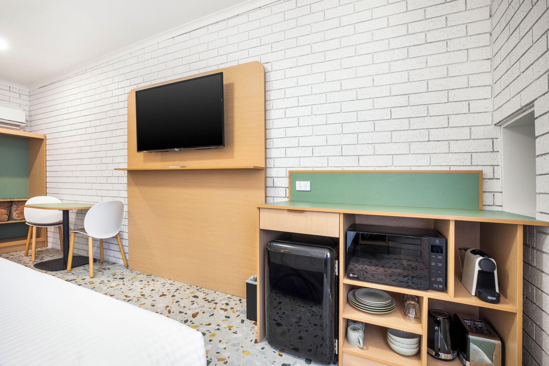 A hotel room with a bed, a TV mounted on the wall, a small kitchenette, and a seating area with a chair and a table.