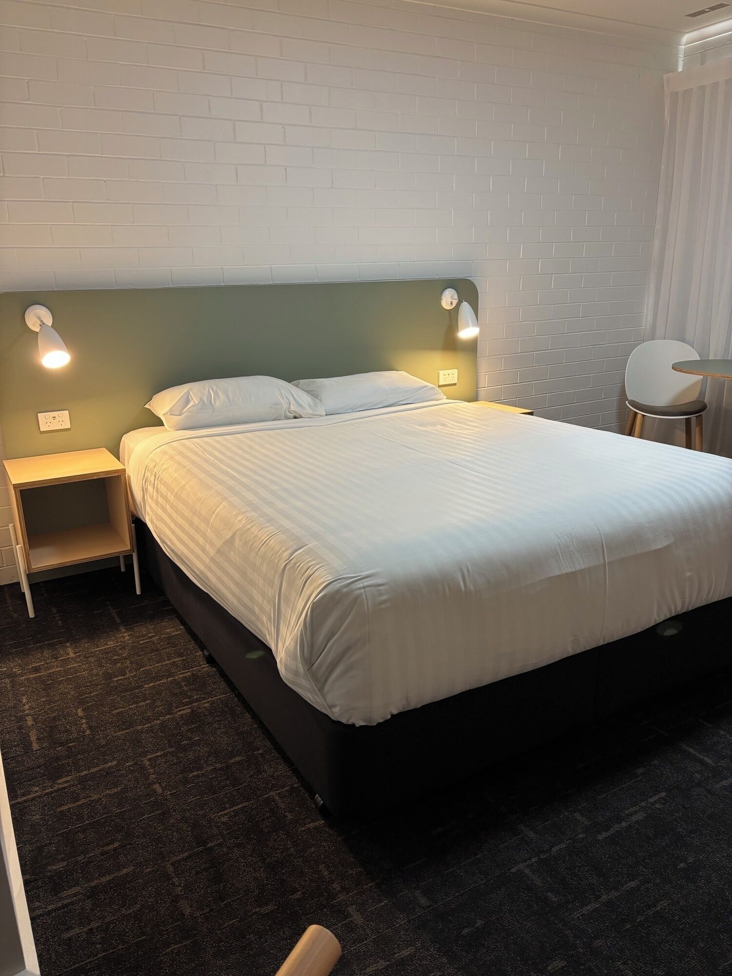 A neatly made bed with white linens, a headboard with a light green panel, two bedside lamps, a small wooden nightstand, and a chair with a round table.