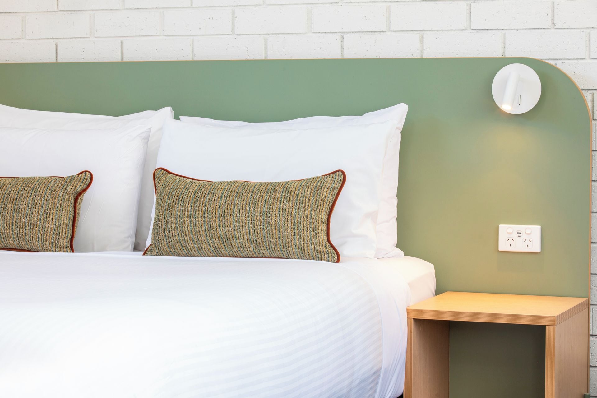 Una cama con ropa blanca y dos almohadas, una mesita de noche y un enchufe en la pared.