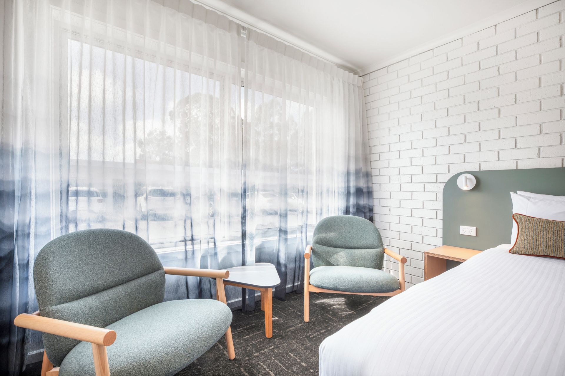 A hotel room with a bed, two armchairs, a small table, and a white brick wall.