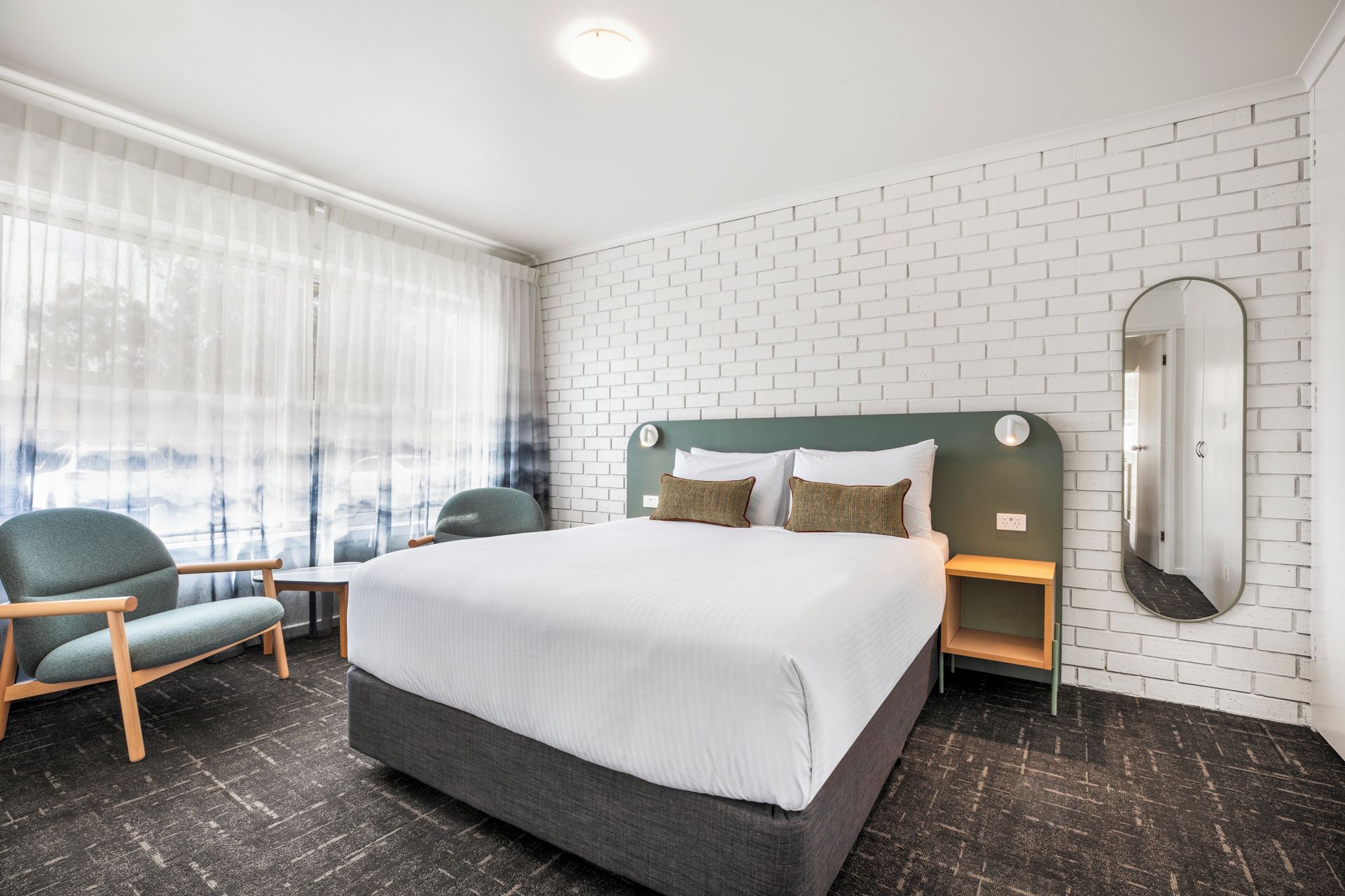 Ein Hotelzimmer mit einem großen Bett, zwei Nachttischen, einem Sessel, einem Fenster mit durchsichtigen Vorhängen und einer Wand aus weißen Ziegeln.