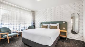 Ein Hotelzimmer mit einem großen Bett, zwei Nachttischen, einem Sessel, einem Fenster mit durchsichtigen Vorhängen und einer Wand aus weißen Ziegeln.
