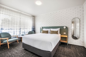 Habitación de hotel con una cama grande, dos mesitas de noche, una silla, una ventana con cortinas traslúcidas y una pared de ladrillo blanco.