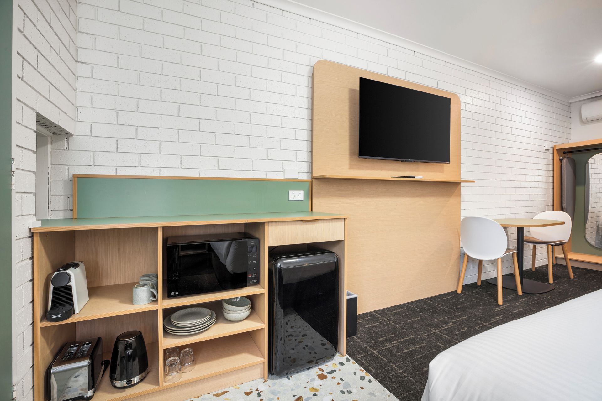 Habitación de hotel con televisor de pantalla plana en la pared, kitchenette con microondas, tostadora y cafetera, una mesita con una silla y una cama con cubrecama blanco.