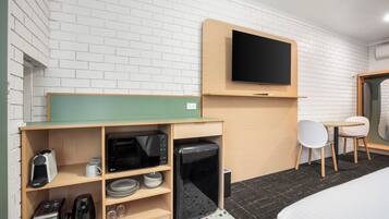 Habitación de hotel con televisor de pantalla plana en la pared, kitchenette con microondas, tostadora y cafetera, una mesita con una silla y una cama con cubrecama blanco.