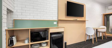 A hotel room with a flat-screen TV mounted on the wall, a kitchenette with a microwave, toaster, and coffee maker, a small table with a chair, and a bed with a white comforter.