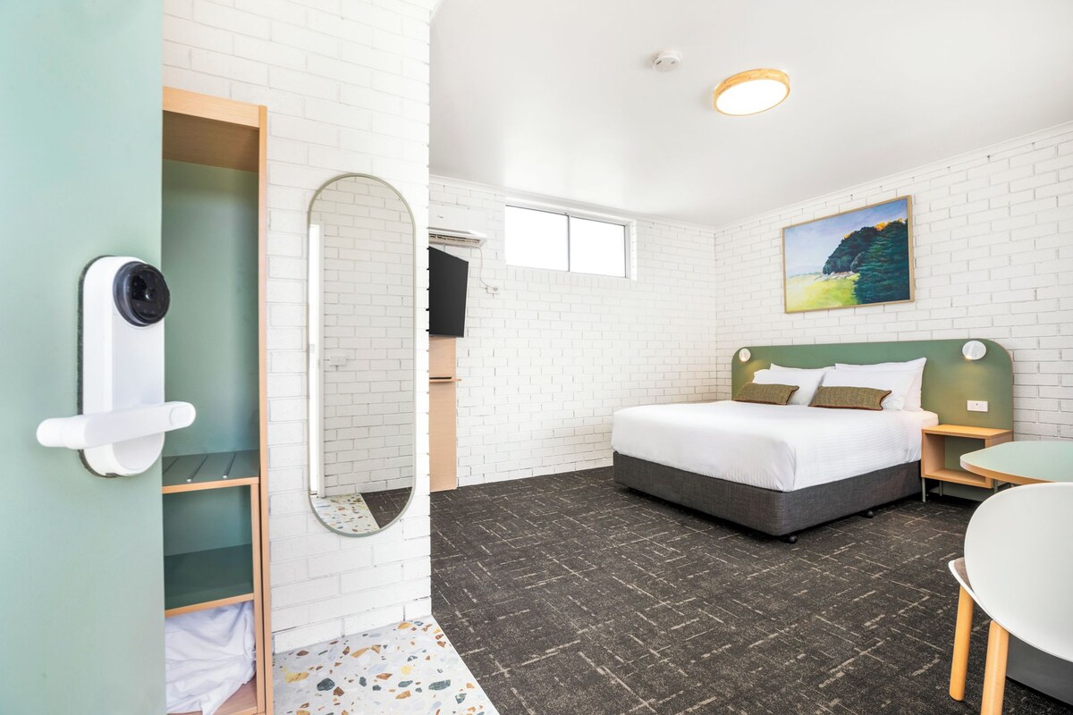 Habitación de hotel con cama, escritorio, silla, espejo, puerta y un cuadro en la pared.