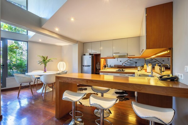 A modern kitchen with a central island, bar stools, and a dining area with a table and chairs. - Petitenget 501 (Keroboken Kelod)