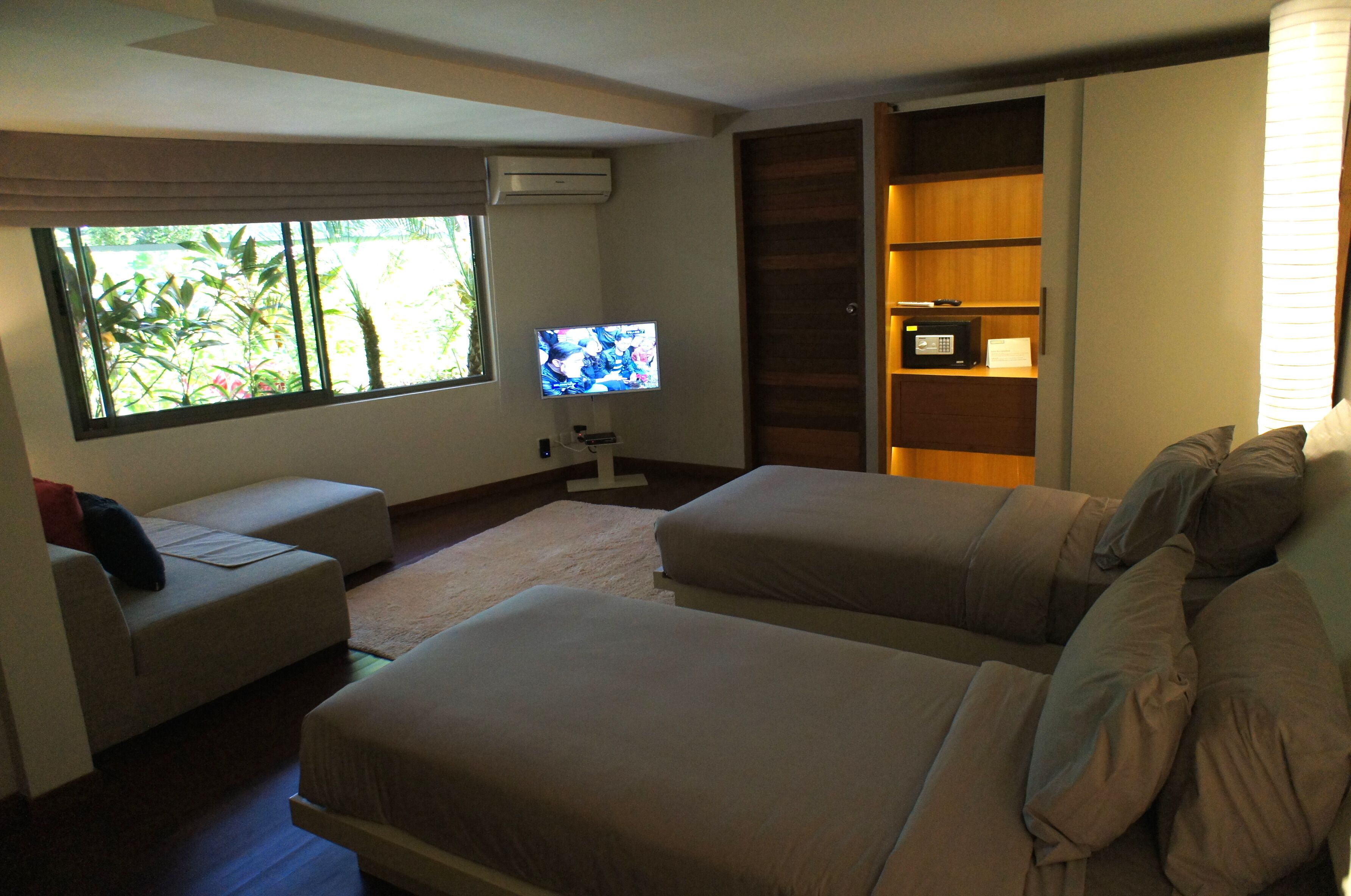 A hotel room with two beds, a TV, a small desk, and a window with a view of greenery.