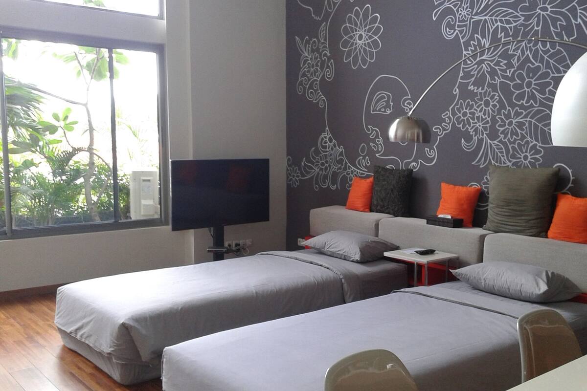 A modern hotel room with two beds, a TV, and a large window with a view of greenery.