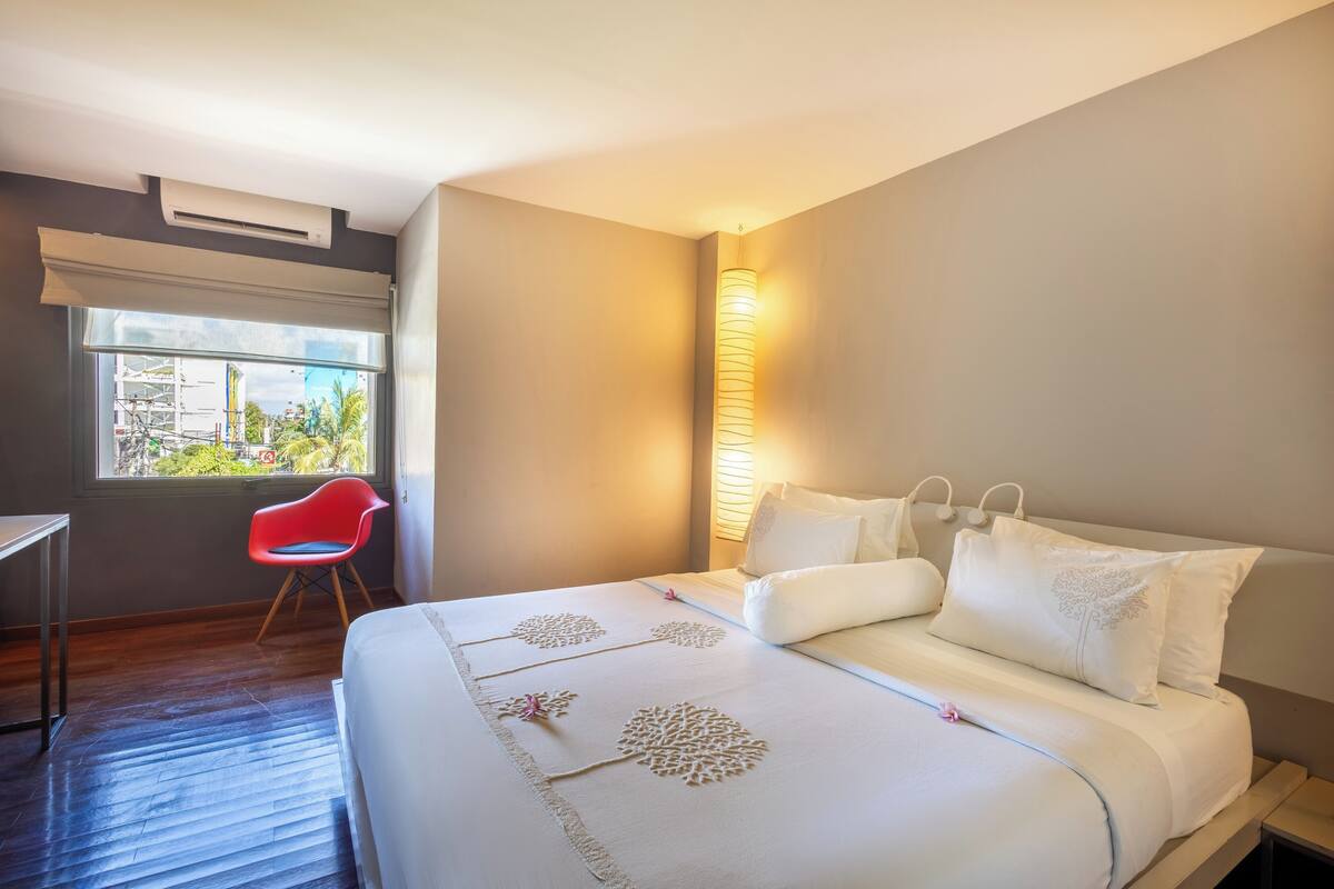 A hotel room with a bed, a red chair, a desk, and a window with a view of buildings and trees.