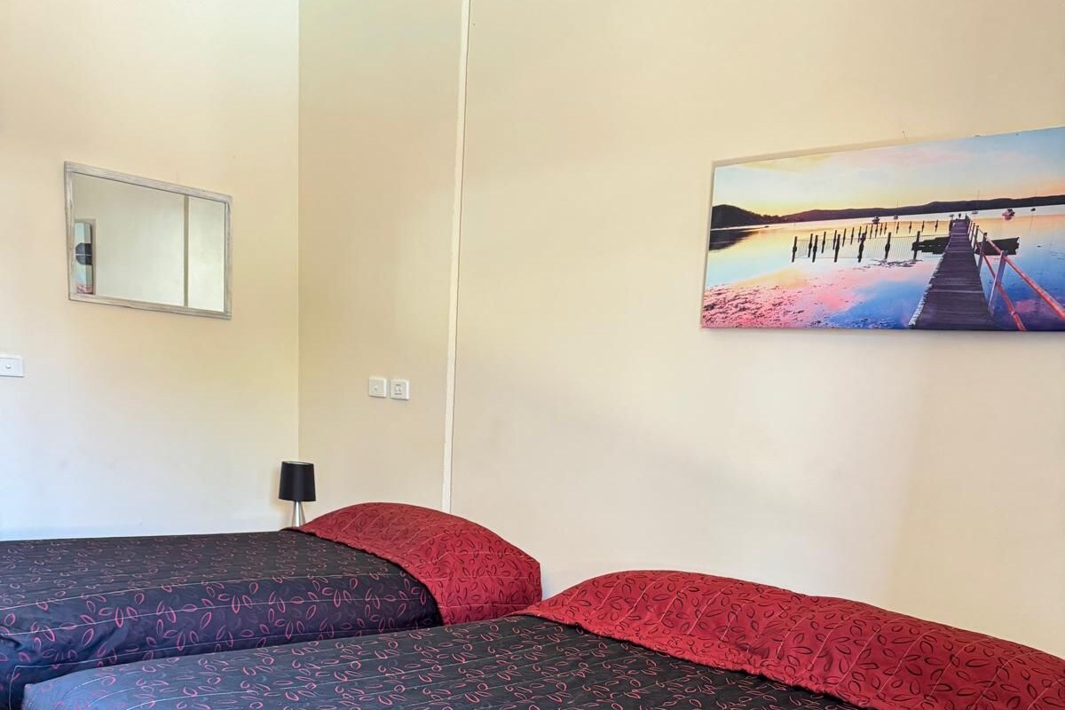 A room with two beds, each with a patterned bedspread, a painting of a sunset on the wall, and a small table with a lamp.