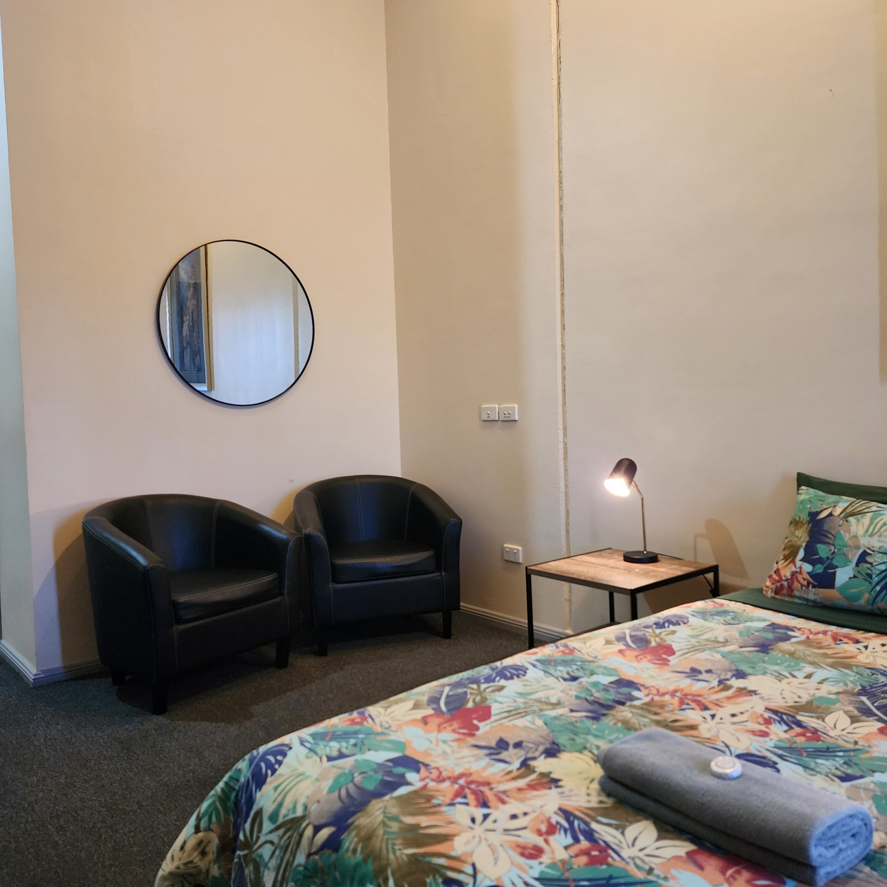 A hotel room with a bed, two chairs, a round mirror, a small table with a lamp, and a floral-patterned bedspread.