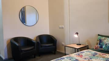 A hotel room with a bed, two chairs, a round mirror, a small table with a lamp, and a floral-patterned bedspread.