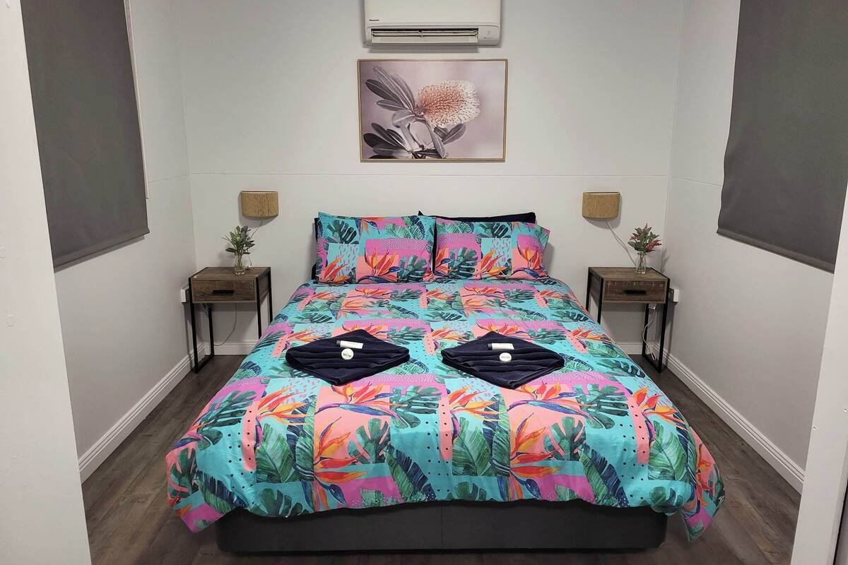 A bedroom with a bed, two bedside tables, an air conditioner, and a wall-mounted lamp.