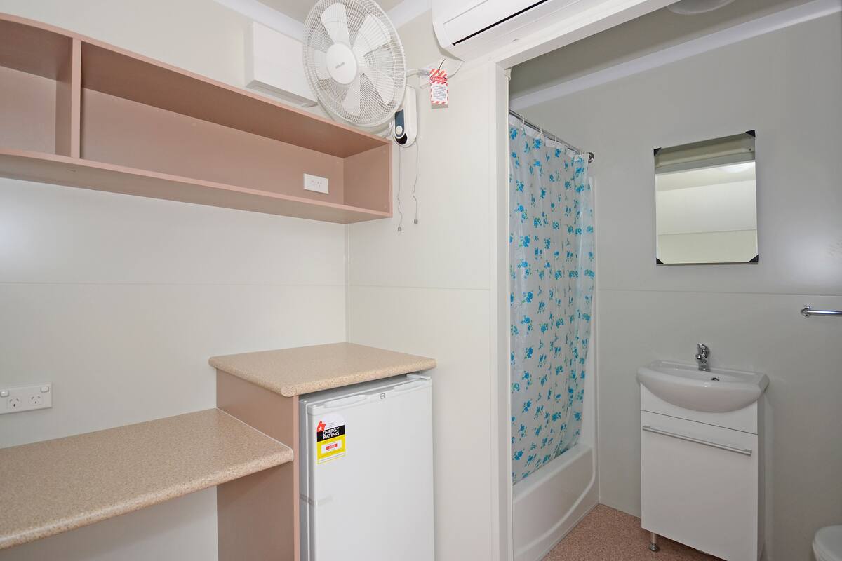 A small, compact bathroom with a white sink, a mirror, a shower with a floral curtain, a small refrigerator, and a ceiling fan.