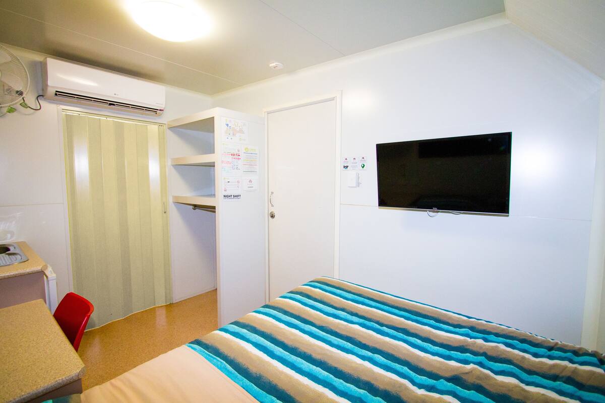 A small, clean room with a bed, a desk, a chair, an air conditioner, a door, and a TV mounted on the wall.