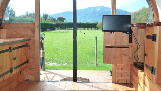 Un interno in legno con un'ampia finestra, una TV a schermo piatto montata a parete e vista su un prato verde con montagne sullo sfondo.