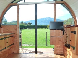 A wooden interior with a large window, a flat-screen TV mounted on the wall, and a view of a grassy field with mountains in the background. - Wacky Stays (Kaikoura)