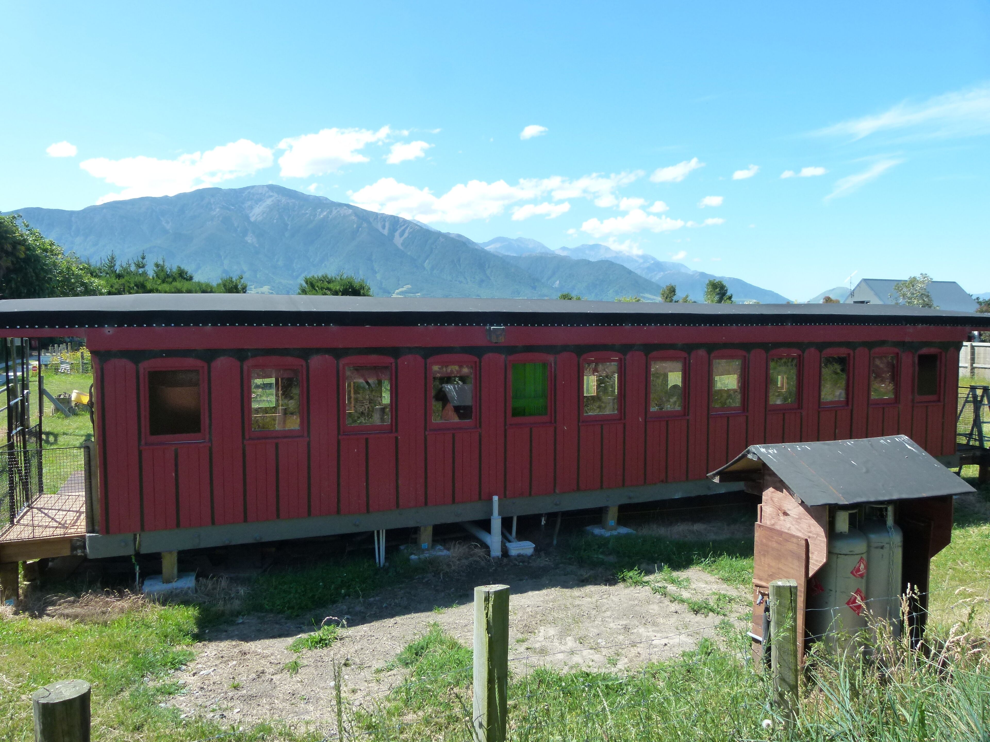 A red train car with multiple windows, set in a rural area with mountains in the background.