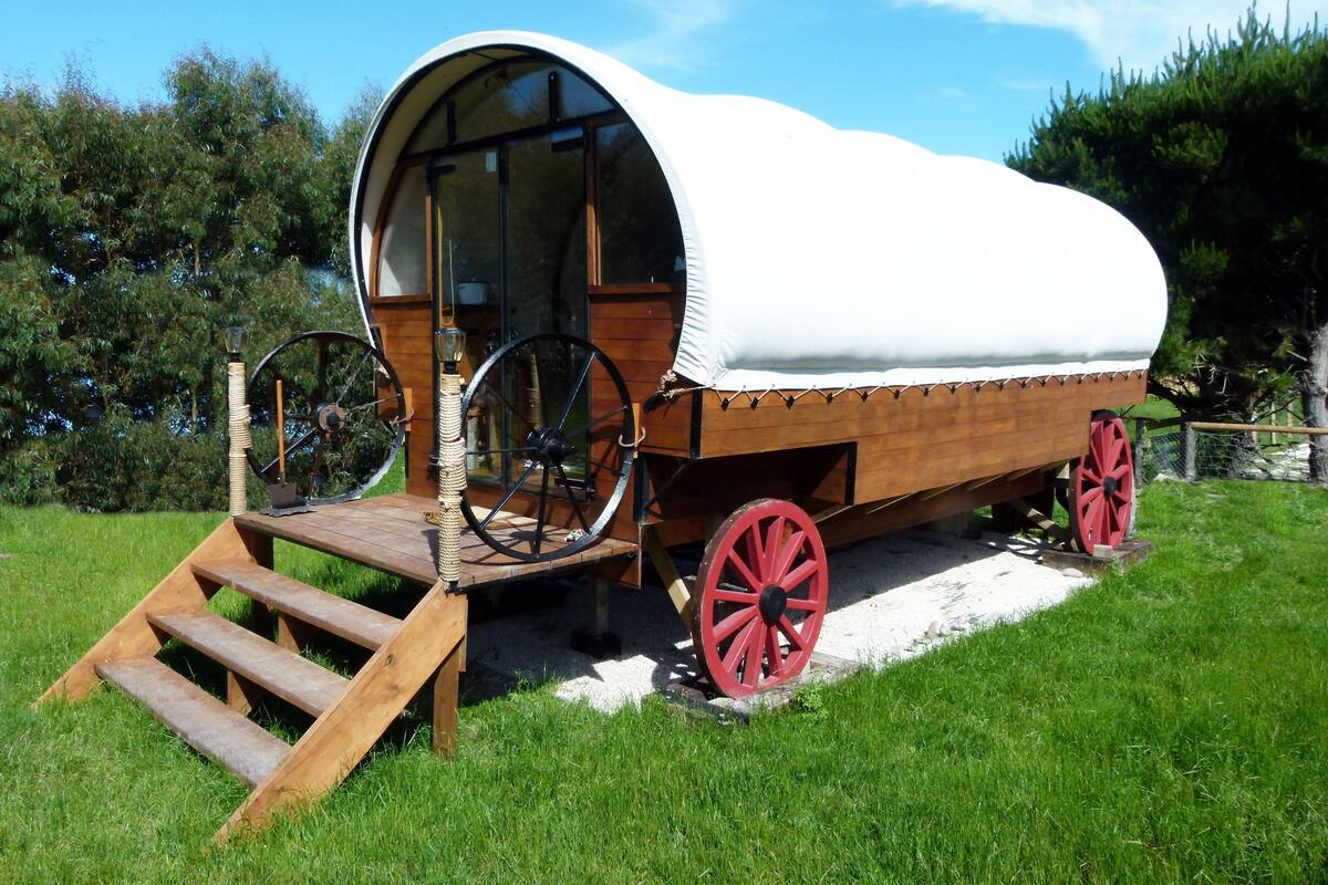 A covered wagon with a wooden structure, a wooden ramp, and red wheels.