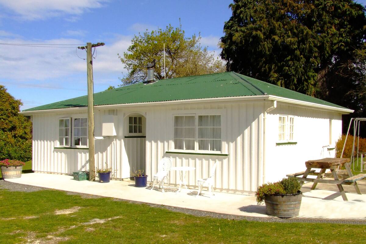 A white building with a green roof, a porch with a bench, and potted plants.