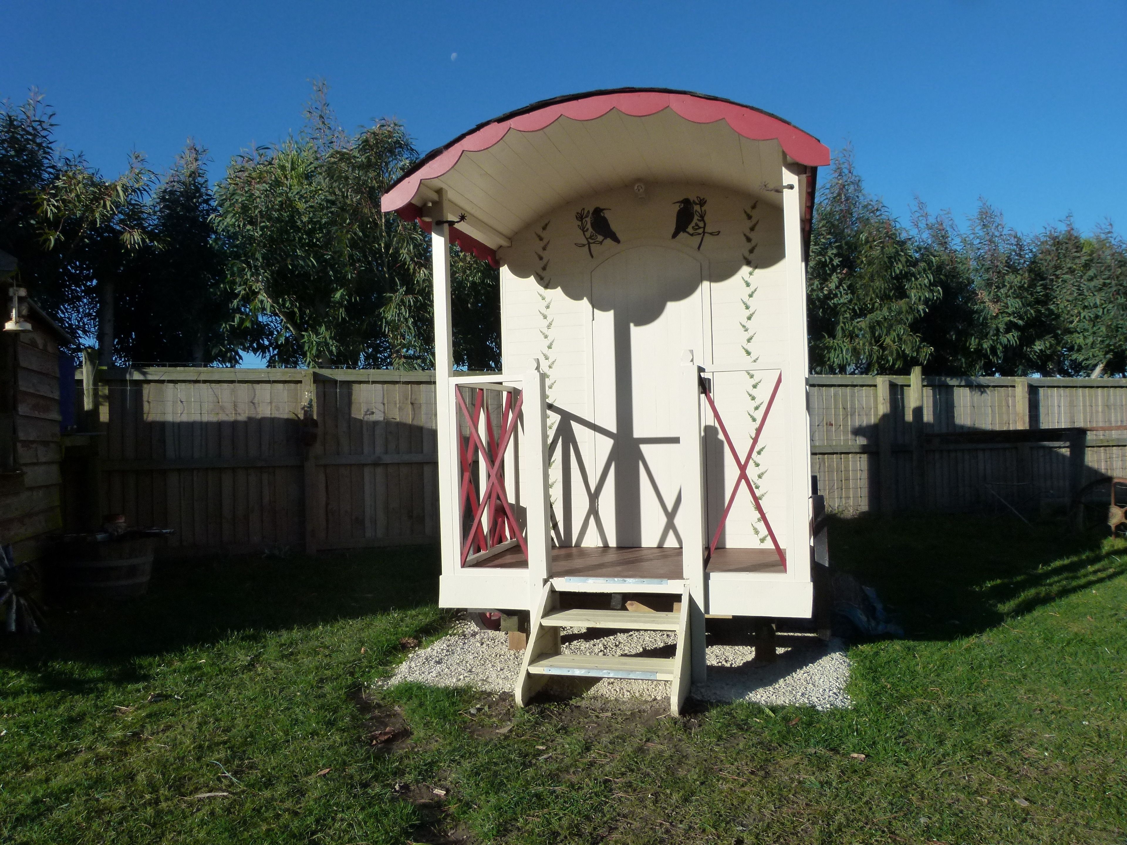 A small, white gazebo with a red roof and decorative bird motifs. It has a wooden bench and a small door with a red cross design.