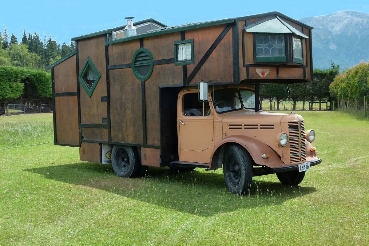 A vintage camper van with a small cabin on top, parked in a grassy field with mountains in the background.
