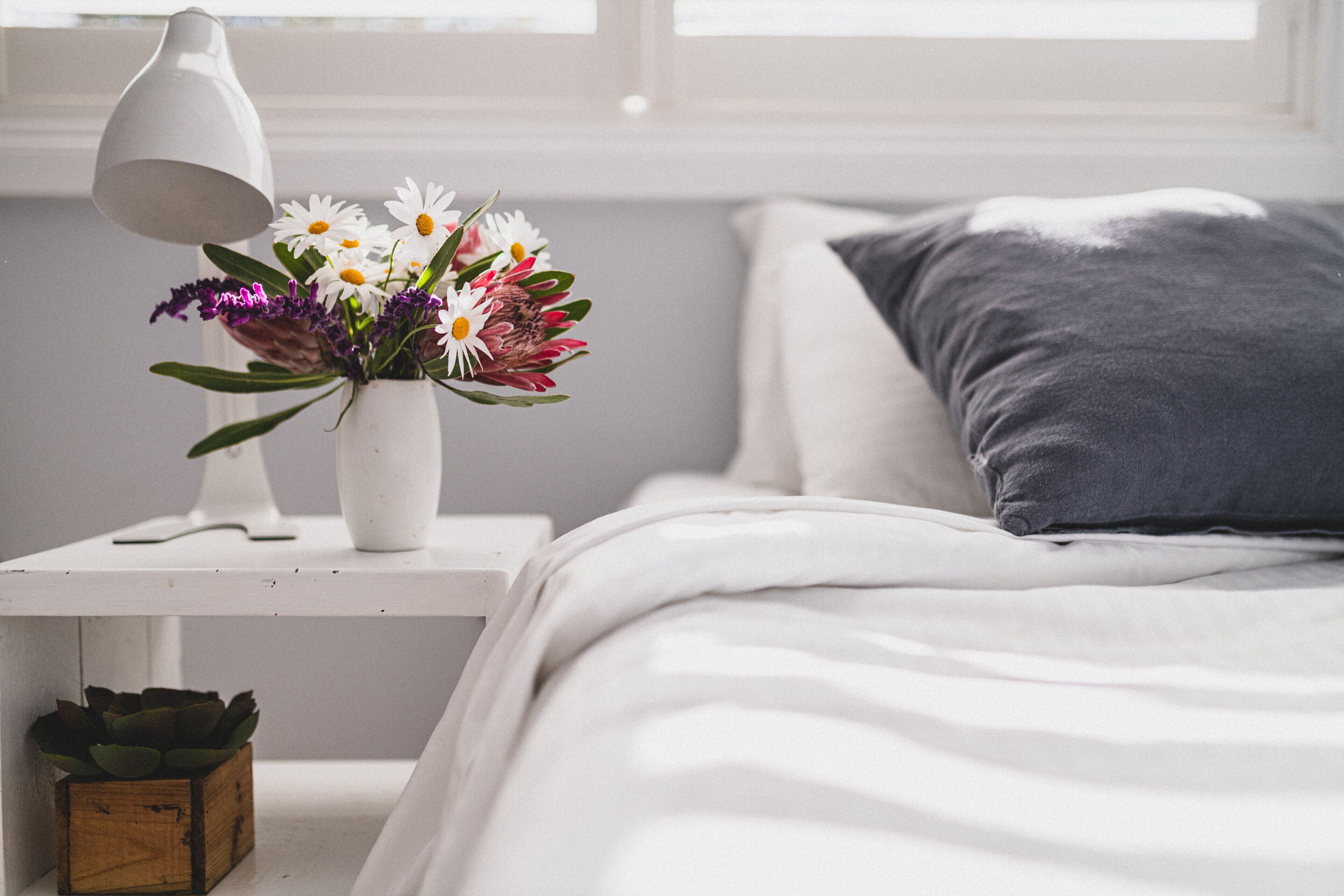 Un lit bien fait, avec un oreiller gris, une table de chevet blanche sur laquelle se trouve une lampe blanche et un vase de fleurs.