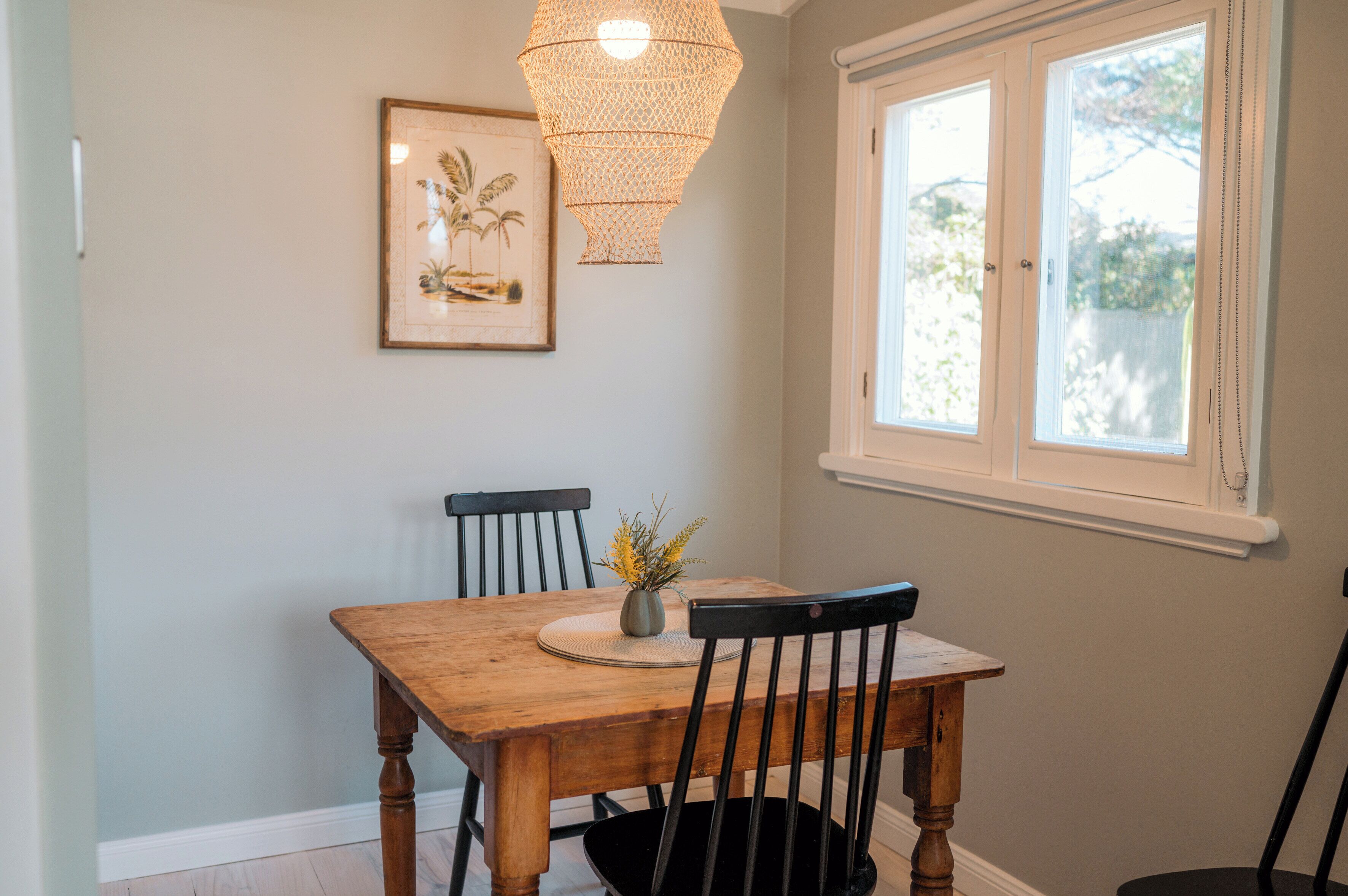 Un coin repas avec une table en bois, deux chaises, un vase avec des fleurs, un tableau encadré au mur et une fenêtre donnant sur des arbres.