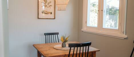 Un coin repas avec une table en bois, deux chaises, un vase avec des fleurs, un tableau encadré au mur et une fenêtre donnant sur des arbres.