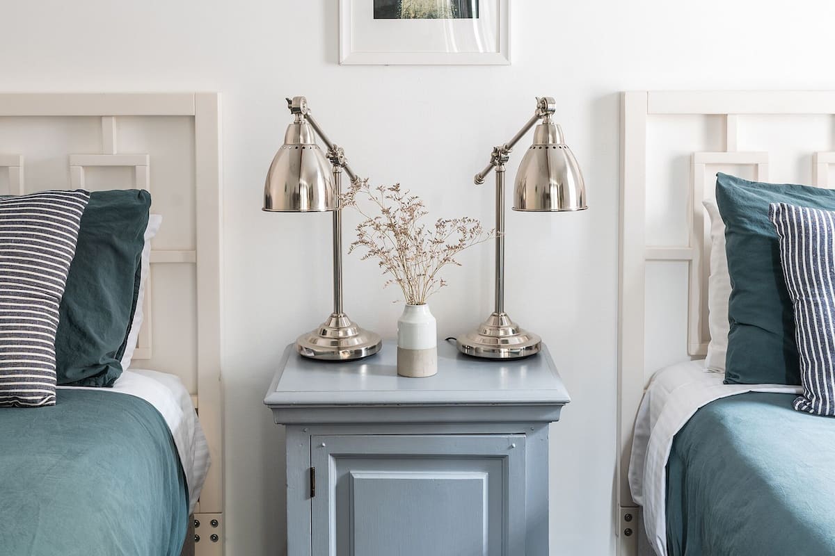 A bedroom with two beds, a nightstand with lamps, a framed picture on the wall, and a small cabinet.