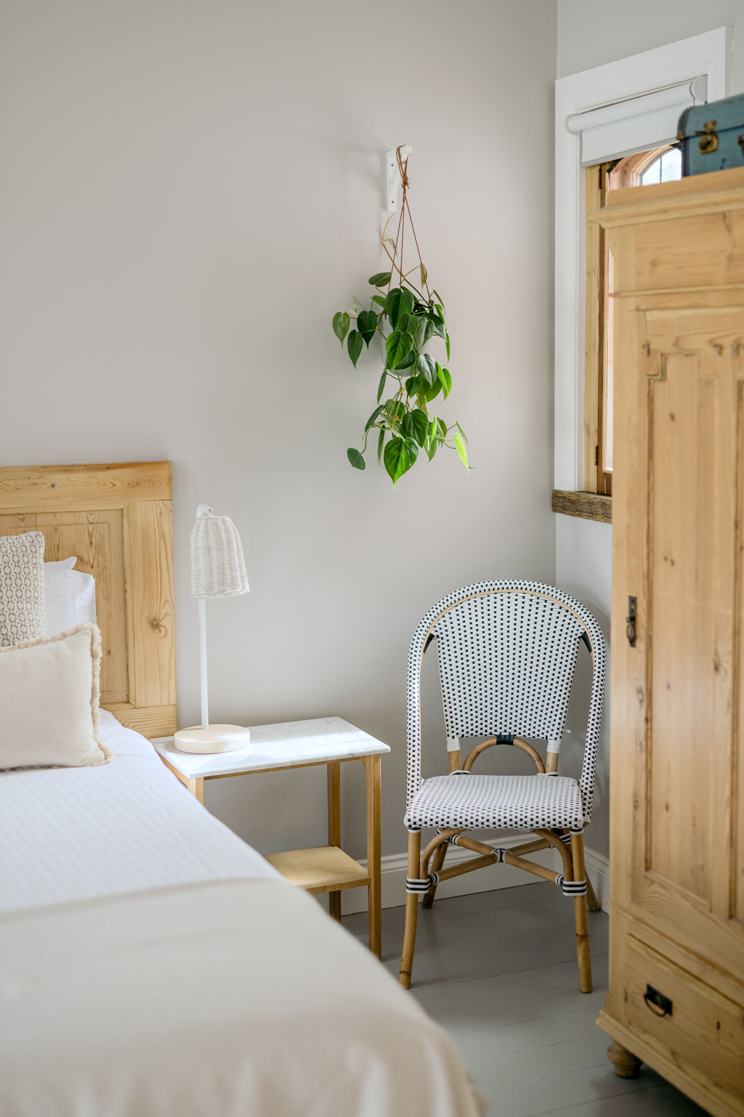 A bedroom with a bed, bedside table, chair, wardrobe, and a hanging plant.
