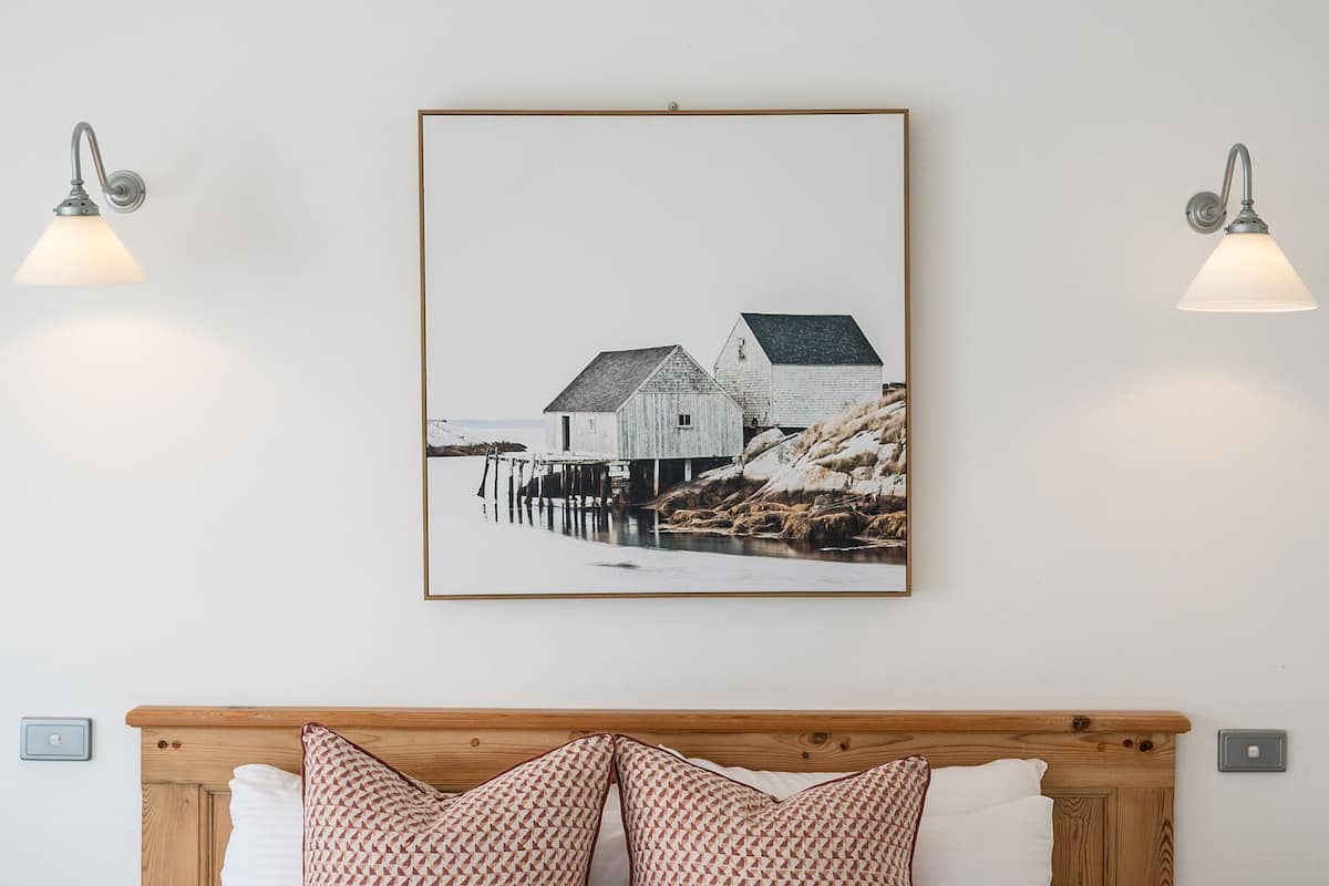 A bed with white bedding and patterned pillows, a framed picture of a coastal scene, and two wall-mounted lamps.