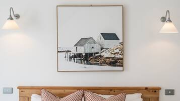 A bed with white bedding and patterned pillows, a framed picture of a coastal scene, and two wall-mounted lamps.